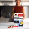 Das Produkt Doppelherz Schwarzkümmelöl 1800 auf einem Tisch, daneben liegen die Kapseln.